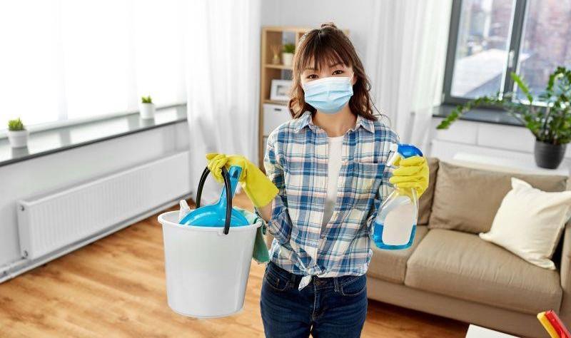 young woman wearing a mask holding a bucket full of spray bottles and chemical bottles