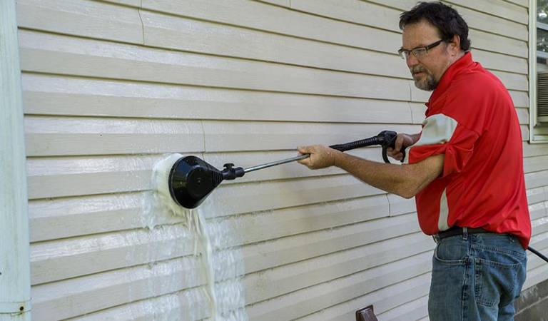 Man in red t-shirt and blue jeans scrubbing exterior shutter with a tick