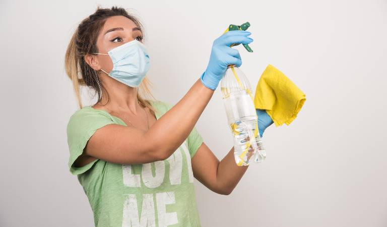 Woman in mask and blue gloves holding a bottle in her hand and a yellow cloth in her second hand.