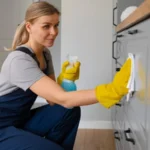 Woman in uniform and yellow gloves cleaning cabinets inside a kitchen.