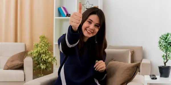 Smiling young pretty caucasian woman sitting on armchair in designed living room looking and showing thumb up