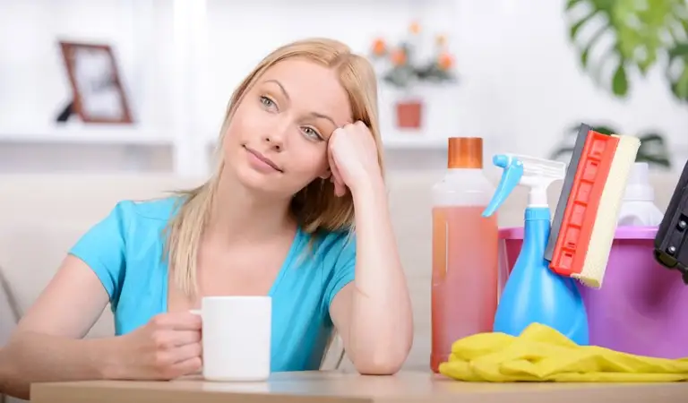 Lady in blue dress drinking coffee with cleaning tools on the table.