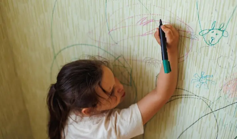 Top view of little naughty girl with dark hair holding green marker in hand drawing pictures on old yellow wallpaper