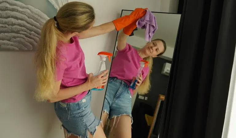 Lady in pink top and blue shorts cleaning mirror with a cloth.