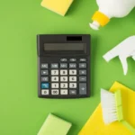 Top view photo of calculator detergent spray gel bottles sponges brush yellow viscose rags and green rubber gloves