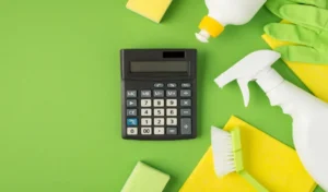 Top view photo of calculator detergent spray gel bottles sponges brush yellow viscose rags and green rubber gloves