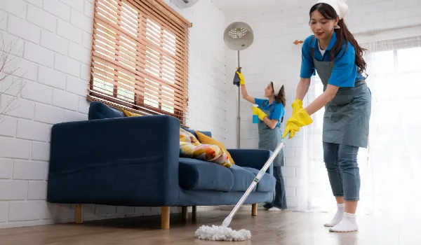 Close up of young woman cleaning floor with mop and 1 dusting in background housework concept