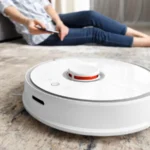 A girl sits on the floor in the living room and controls a robotic vacuum cleaner using a smartphone